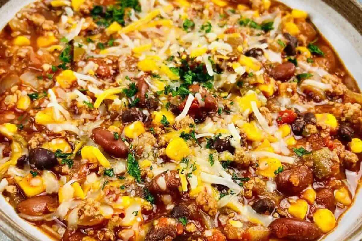 A must-try bowl of chili topped with shredded cheese and chopped herbs, featuring ground meat, beans, corn, tomatoes, and visible spices—a favorite in chili recipes that everyone loves.