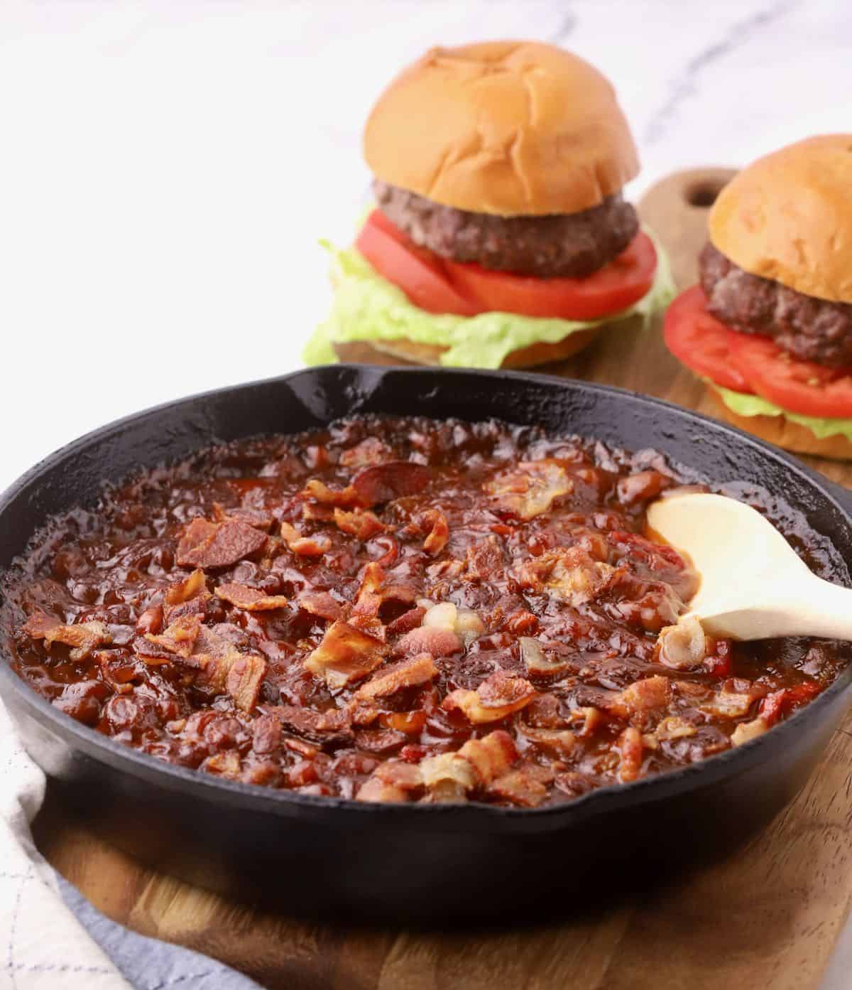 A skillet of baked beans topped with bacon and a serving spoon sits on a wooden board, perfect for potluck dishes. Nearby, two juicy burgers with lettuce and tomato await the crowd that's sure to be recipe asking after tasting this foolproof delight.