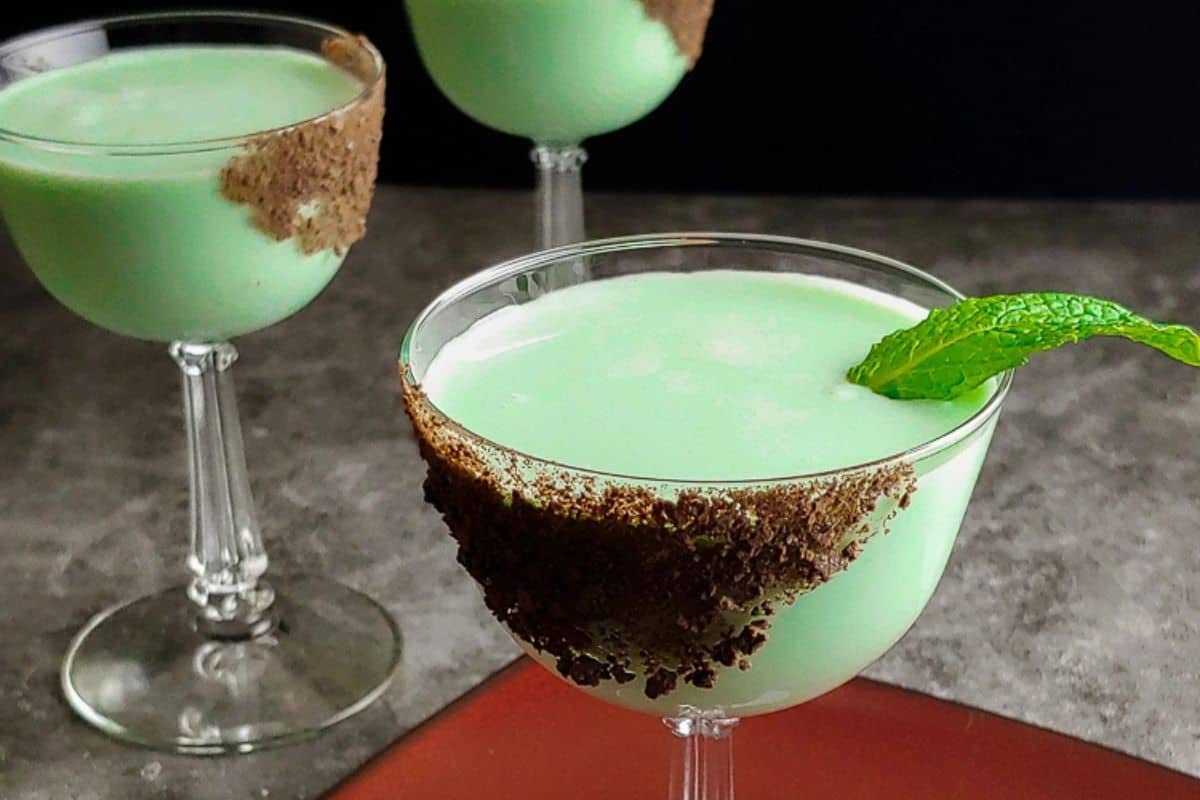 Three must-try green cocktails in stemmed glasses, rimmed with chocolate shavings, embody the Irish spirit. One glass boasts a mint leaf garnish, making them perfect for St. Patrick's Day drinks.