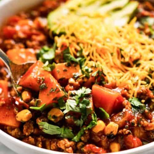 A bowl of must try chili with ground meat, corn, tomatoes, and beans, topped with shredded cheese, avocado slices, and cilantro. A spoon is in the bowl—perfect for fans of popular chili recipes.