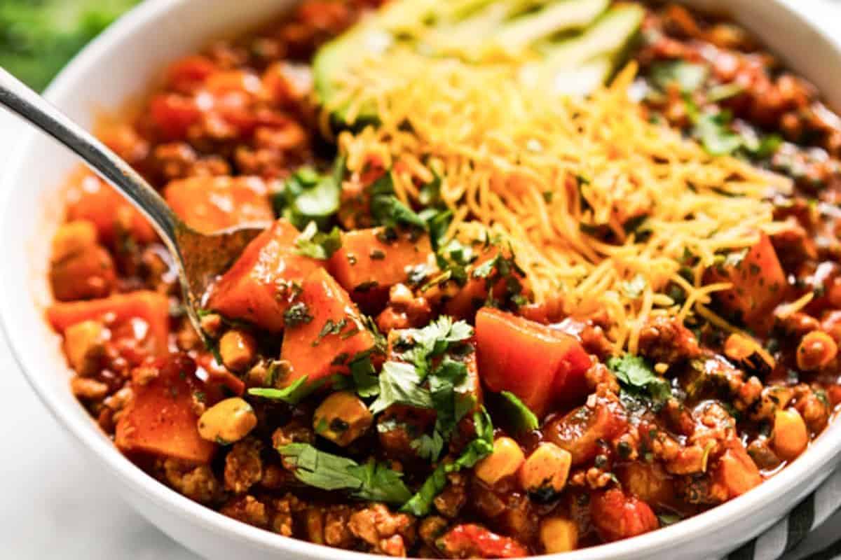 A bowl of must try chili with ground meat, corn, tomatoes, and beans, topped with shredded cheese, avocado slices, and cilantro. A spoon is in the bowl—perfect for fans of popular chili recipes.