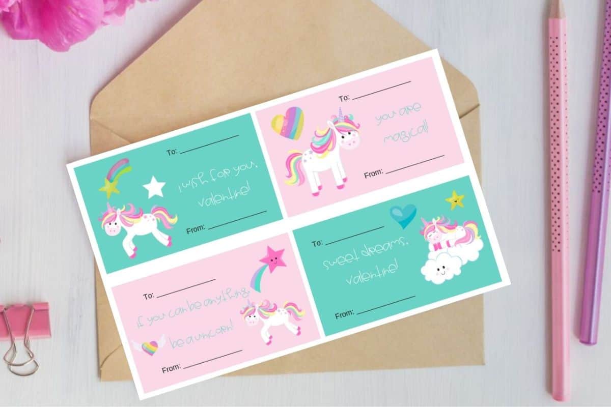 A sheet of four pastel-colored unicorn-themed Printable Valentines with “To” and “From” fields, placed on a brown envelope next to pink stationery items.