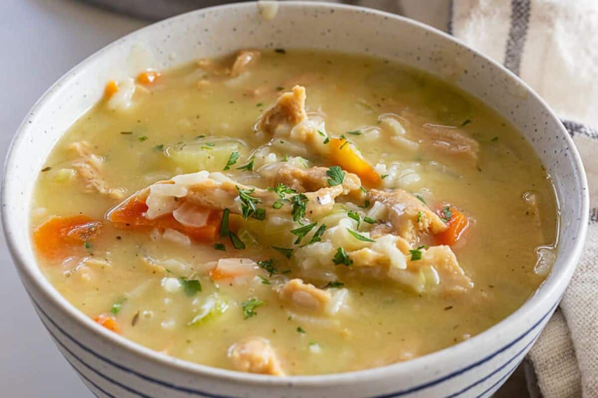 A bowl of creamy chicken and rice soup with chunks of chicken, carrots, celery, and parsley sits next to a striped cloth—one of those comforting soups perfect for cozy nights.