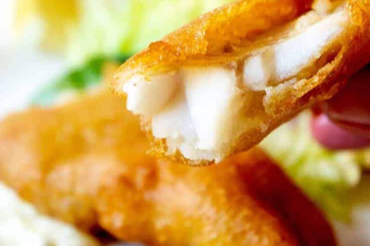 Close-up of a piece of battered and fried fish with white, flaky interior, held in front of a blurred background—perfect for Lent and those seeking easy recipes for classic fish dishes.