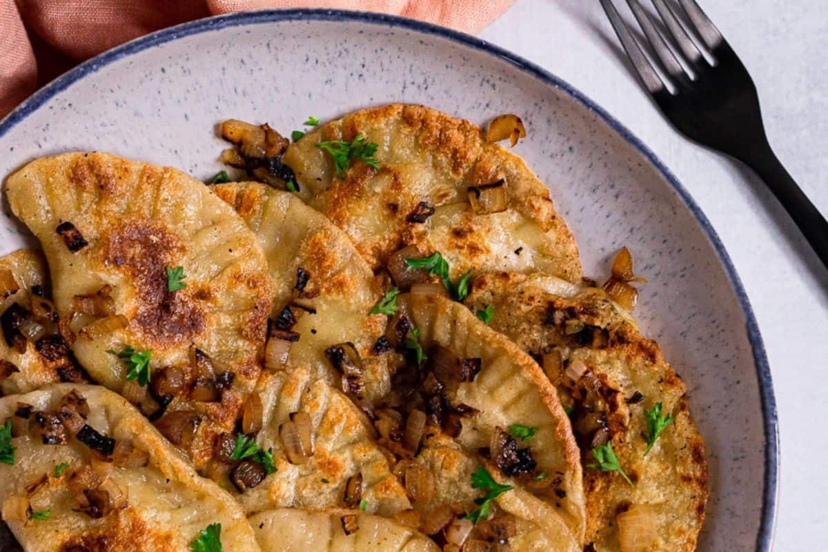 A plate of irresistible pan-fried pierogies with caramelized onions and parsley, next to a black fork and a peach-colored napkin, is sure to satisfy your cravings.