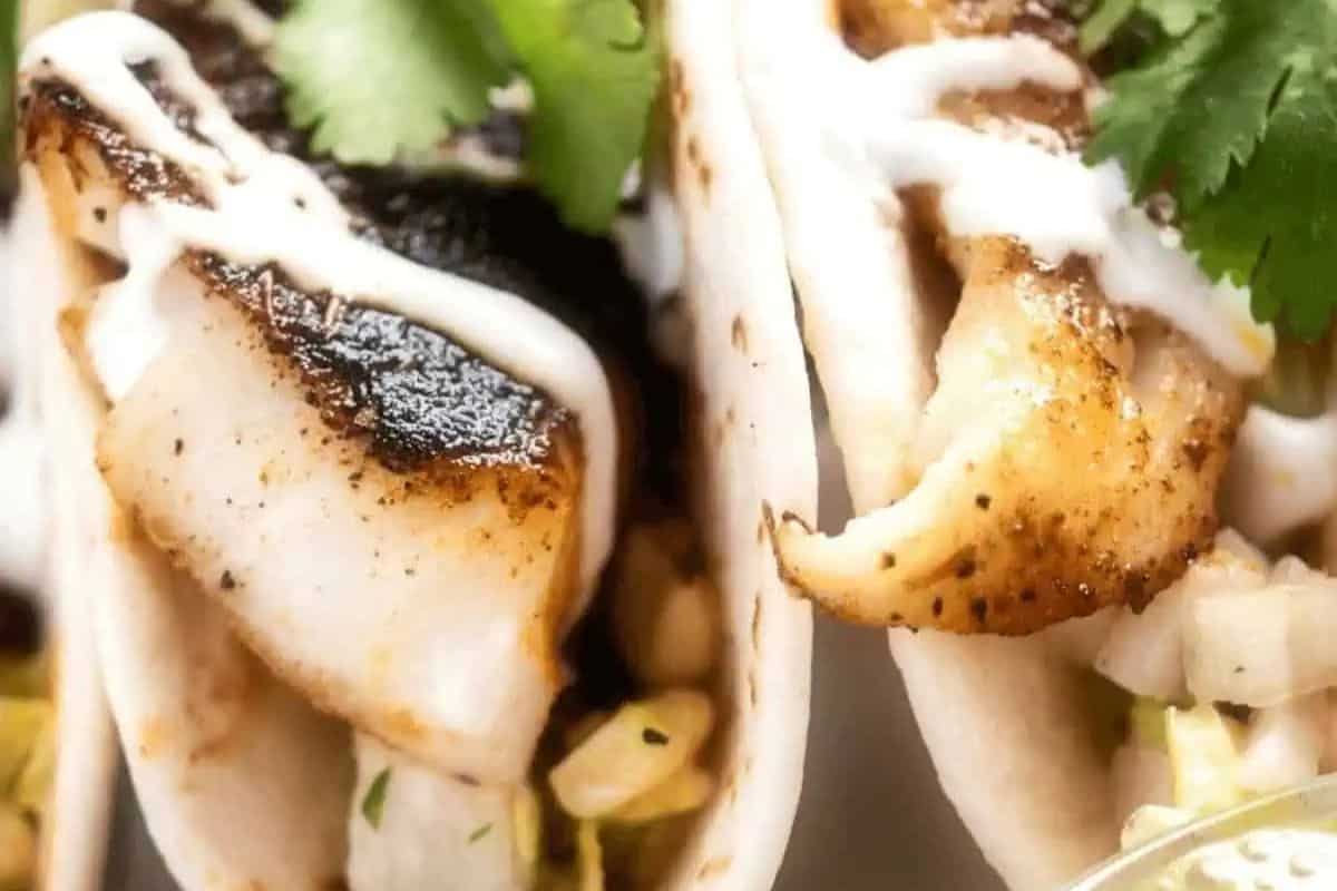 Close-up of two fish tacos—an easy recipe featuring grilled fish, diced onions, lettuce, cilantro, and a drizzle of white sauce in soft tortillas. Perfect for Lent or anyone who loves fresh fish dishes.