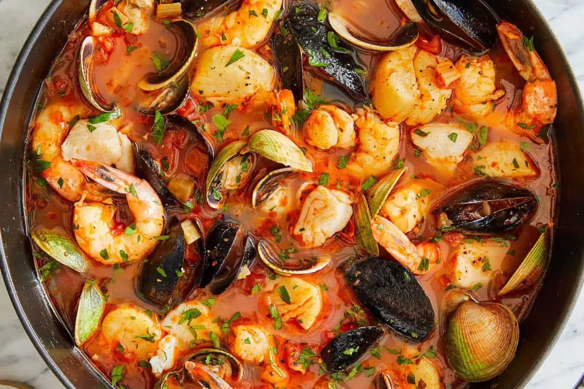 A pan filled with seafood stew—a delightful choice for light meals or Lent recipes—containing shrimp, mussels, clams, and scallops in a tomato-based broth, garnished with chopped herbs.