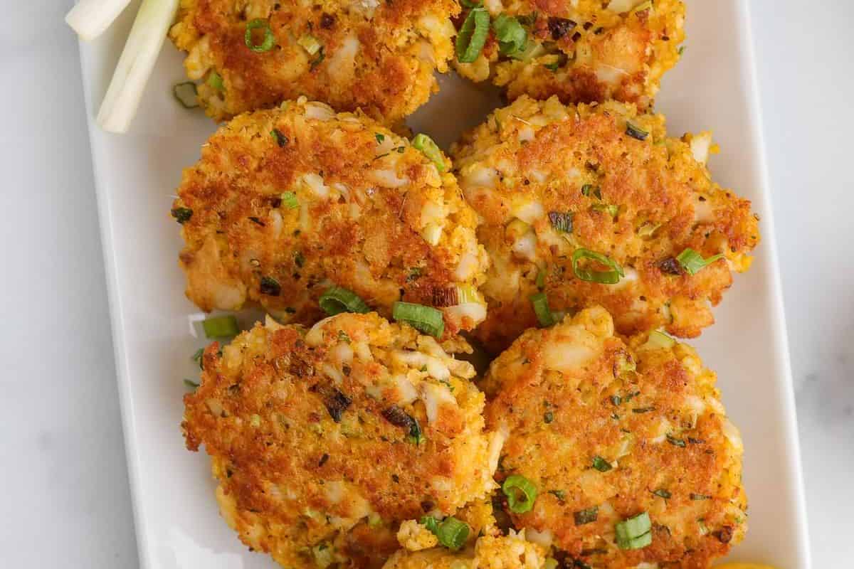 Six golden-brown crab cakes garnished with chopped green onions are arranged on a white rectangular plate—an ideal choice for light meals or those seeking delicious Lent recipes.