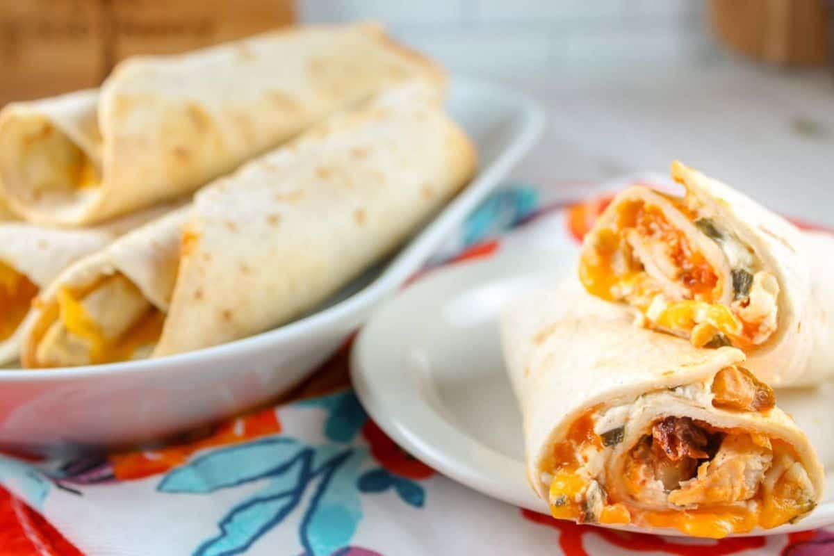 Close-up of rolled tortillas filled with chicken, cheese, and vegetables on a white plate, reminiscent of homemade burritos for a Cinco de Mayo feast, with more on a larger platter in the background.