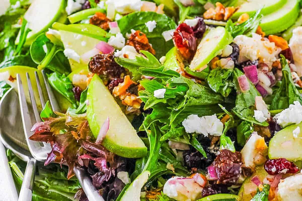 Close-up of a fresh spring salad with mixed greens, sliced green apples, red onions, walnuts, cranberries, and feta cheese—perfectly embodying the essence of seasonal salad recipes. A fork waits invitingly at the side.