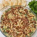 A round glass dish filled with artichoke and spinach dip recipe, topped with chopped pecans and breadcrumbs, placed next to sliced bread and fresh parsley.