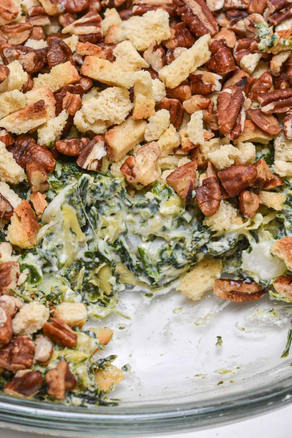 A close-up of a spinach casserole inspired by an Artichoke and Spinach Dip Recipe, topped with chopped pecans and croutons, with a portion removed to reveal the creamy spinach filling.