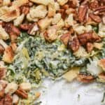 A close-up of a spinach casserole inspired by an Artichoke and Spinach Dip Recipe, topped with chopped pecans and croutons, with a portion removed to reveal the creamy spinach filling.