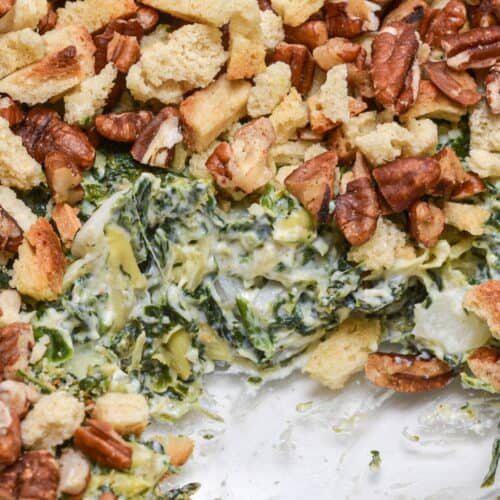 A close-up of a spinach casserole inspired by an Artichoke and Spinach Dip Recipe, topped with chopped pecans and croutons, with a portion removed to reveal the creamy spinach filling.