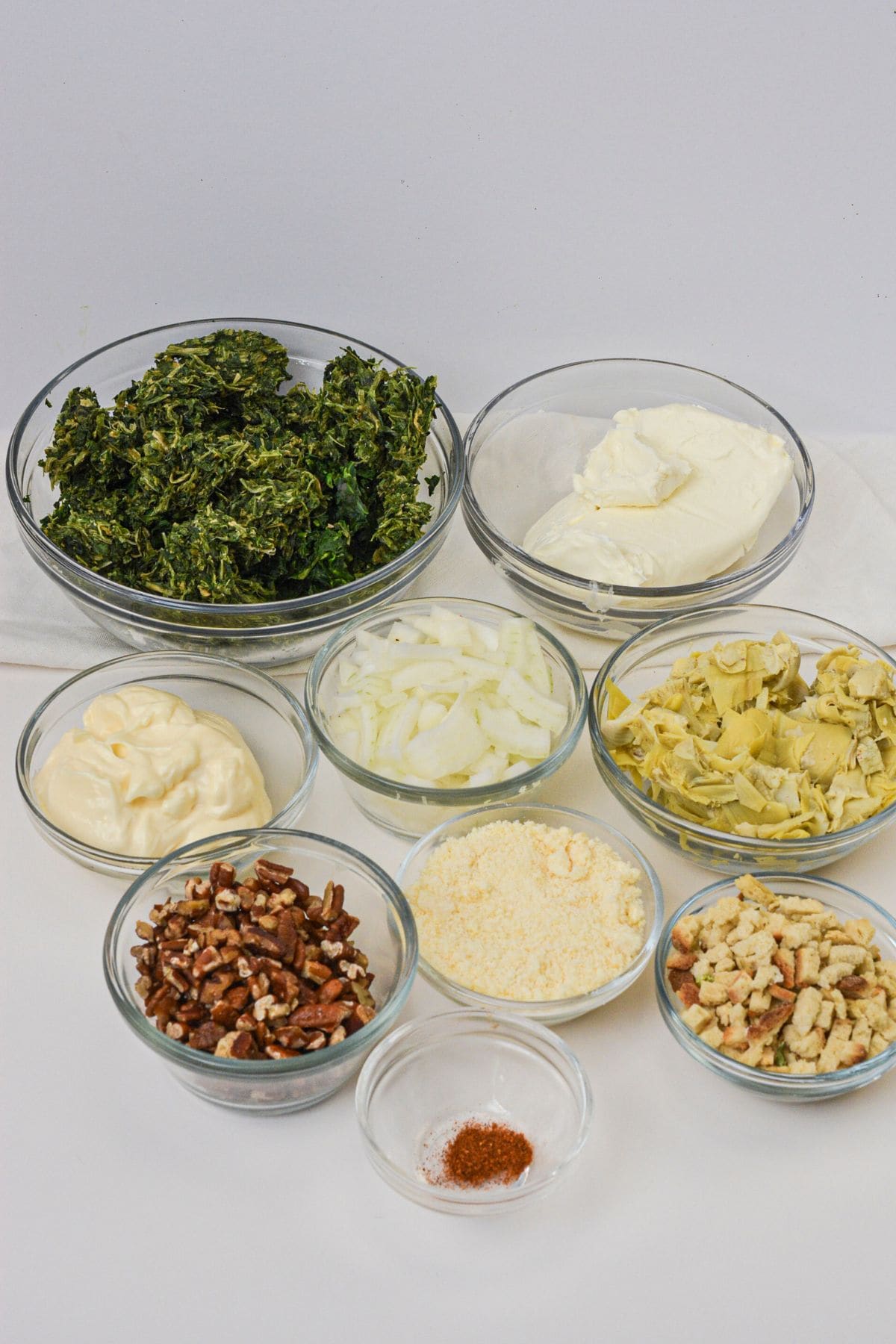 Eight glass bowls filled with chopped kale, cream cheese, mayonnaise, sliced onions, artichokes, pecans, grated cheese, croutons, and spices.