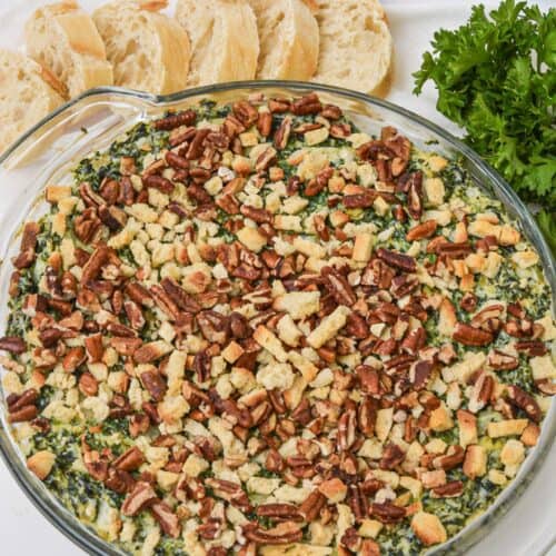 A round glass dish filled with a baked Artichoke and Spinach Dip Recipe, topped with chopped pecans and breadcrumbs, served alongside sliced baguette and fresh parsley.