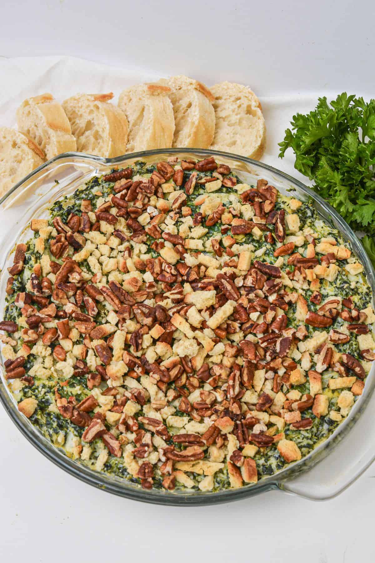 A round glass dish filled with a baked Artichoke and Spinach Dip Recipe, topped with chopped pecans and breadcrumbs, served alongside sliced baguette and fresh parsley.