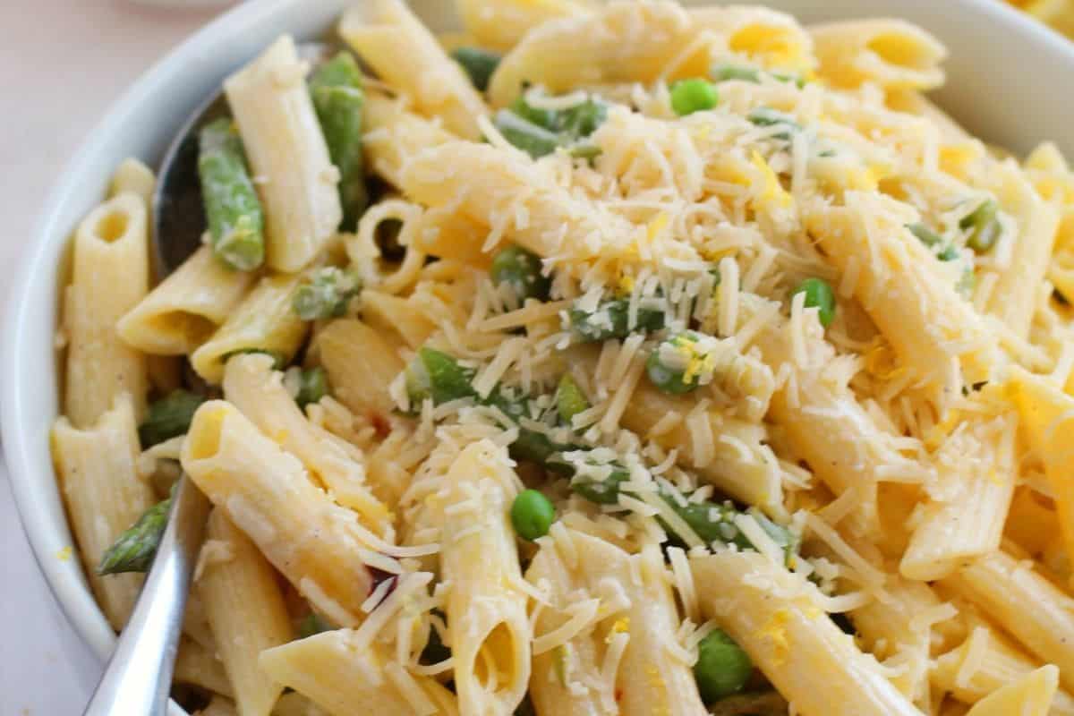 A bowl of penne pasta with asparagus, peas, and grated cheese on top. A spoon is placed in the bowl, making this a must-try spring delight among pasta dishes.
