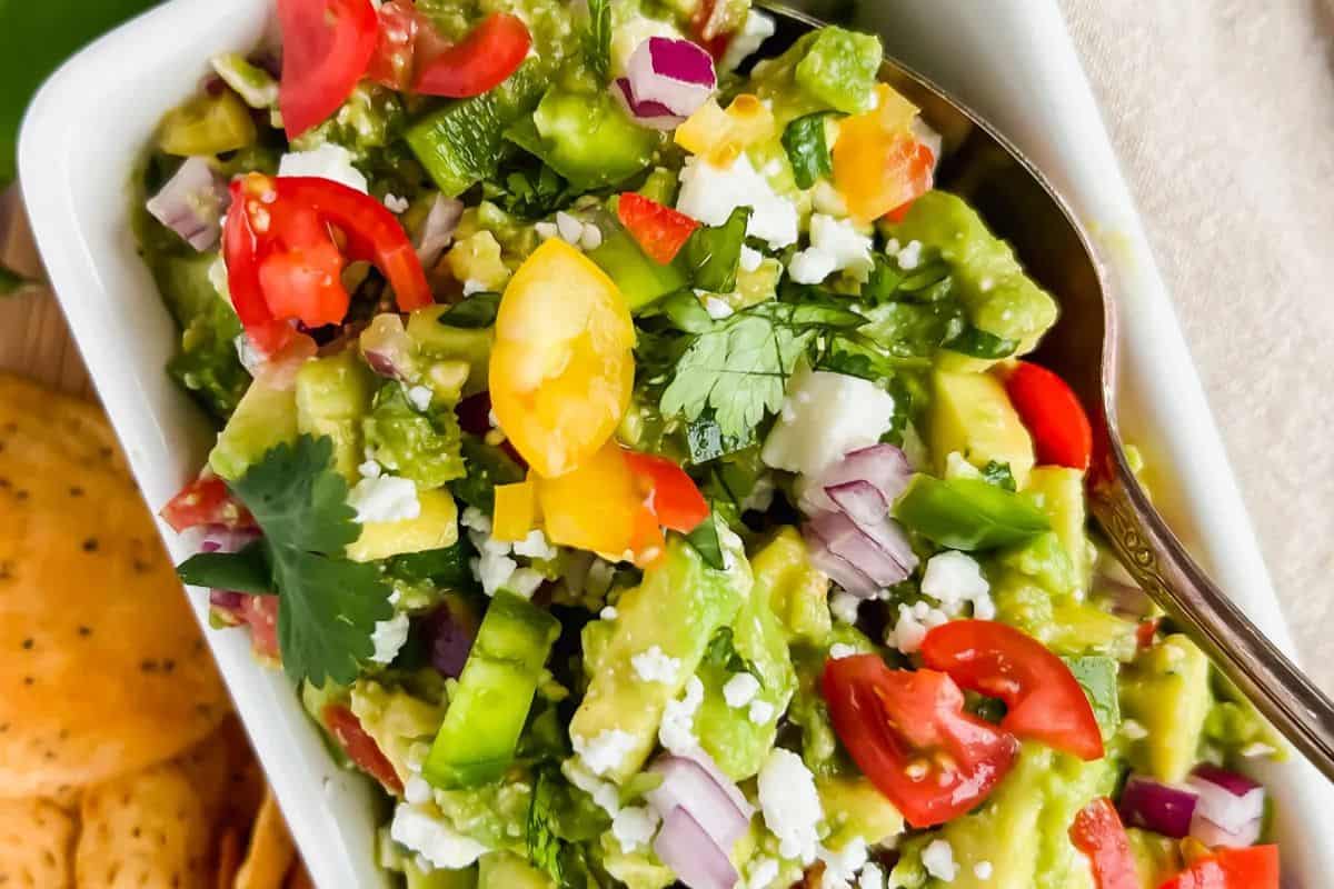 A close-up of a festive party dip: a bowl of guacamole topped with diced tomatoes, red onion, cilantro, and crumbled cheese, complete with a spoon for the perfect Cinco de Mayo celebration.