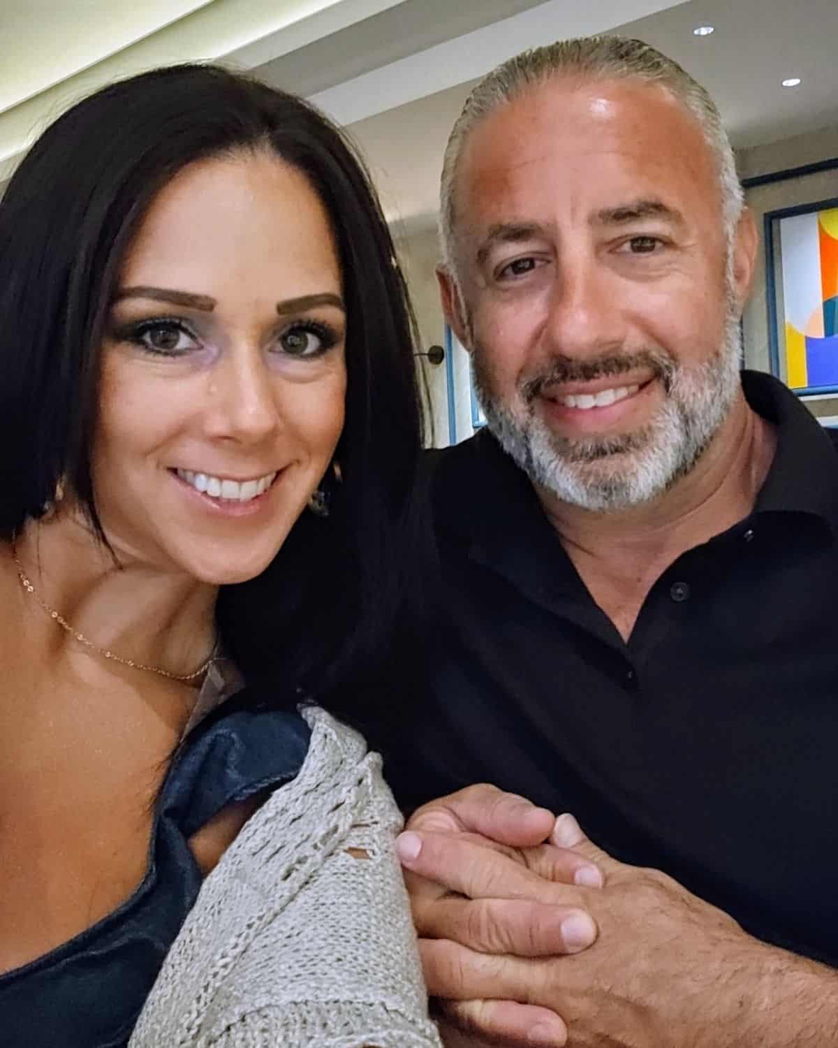 A woman and a man are sitting together, smiling at the camera. She has long dark hair, and he sports short gray hair and a beard. Indoors, with art on the wall behind them, they look like they just finished learning how to make gnocchi in 7 minutes.