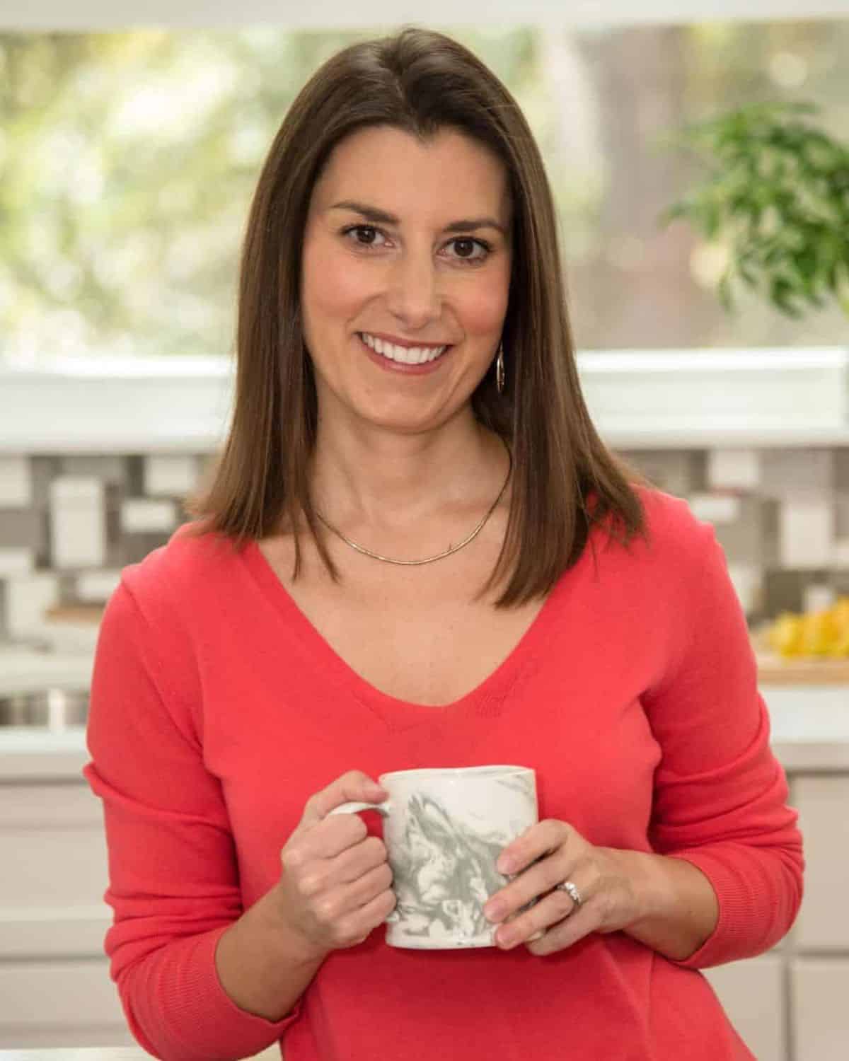 A person with long brown hair in a coral sweater holds a mug, smiling in a kitchen that hints at their passion for diet planning.