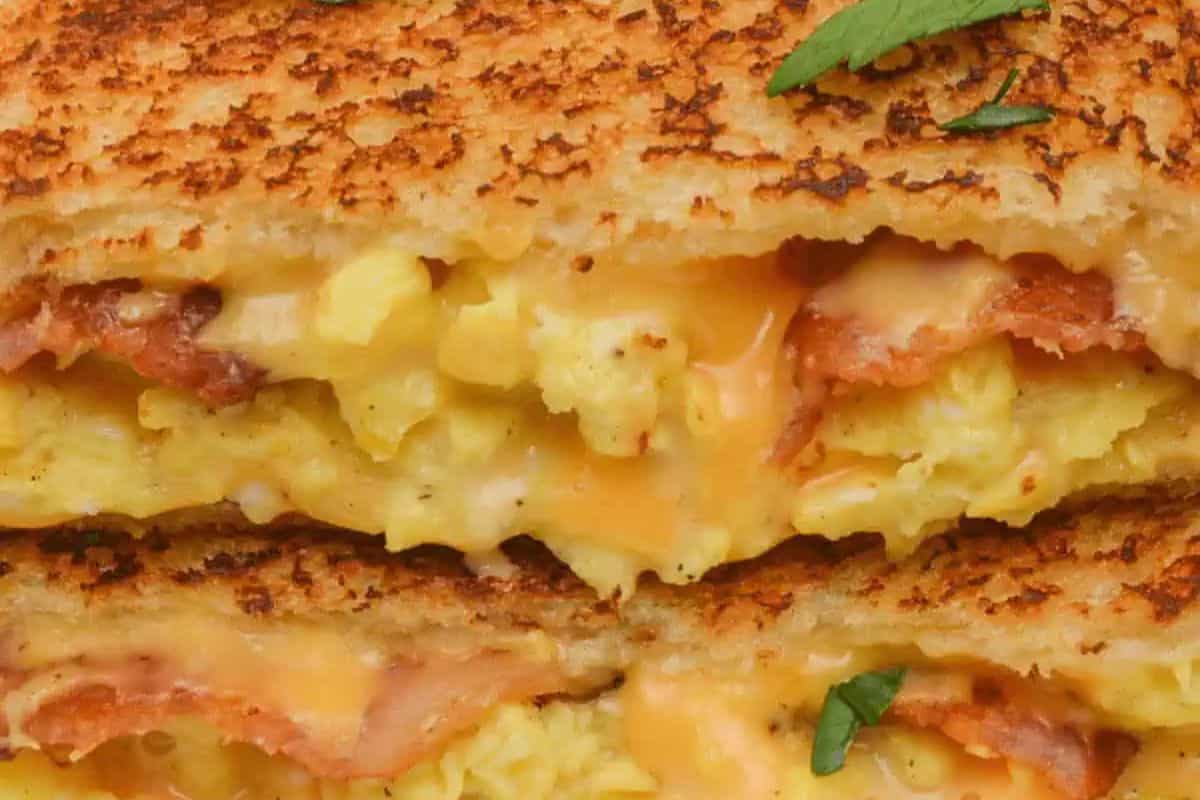 Close-up of a grilled sandwich, perfect for spring brunch recipes, featuring layers of melted cheese, scrambled eggs, and bacon, all garnished with a small green herb leaf on top.