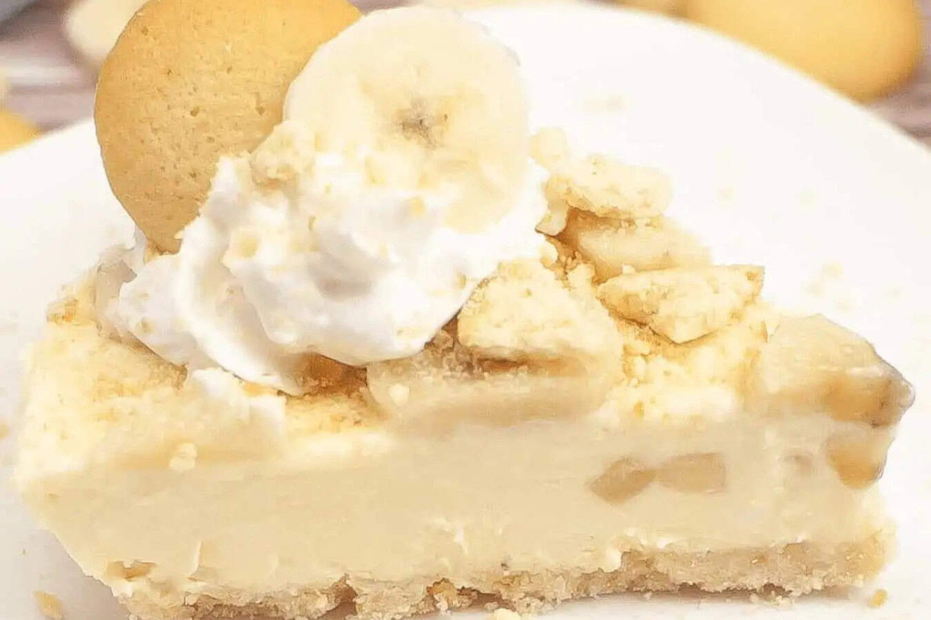 A slice of banana pudding cheesecake, epitomizing beautiful desserts, is elegantly topped with whipped cream, banana slices, crumbled cookies, and crowned with a whole cookie on a white plate.
