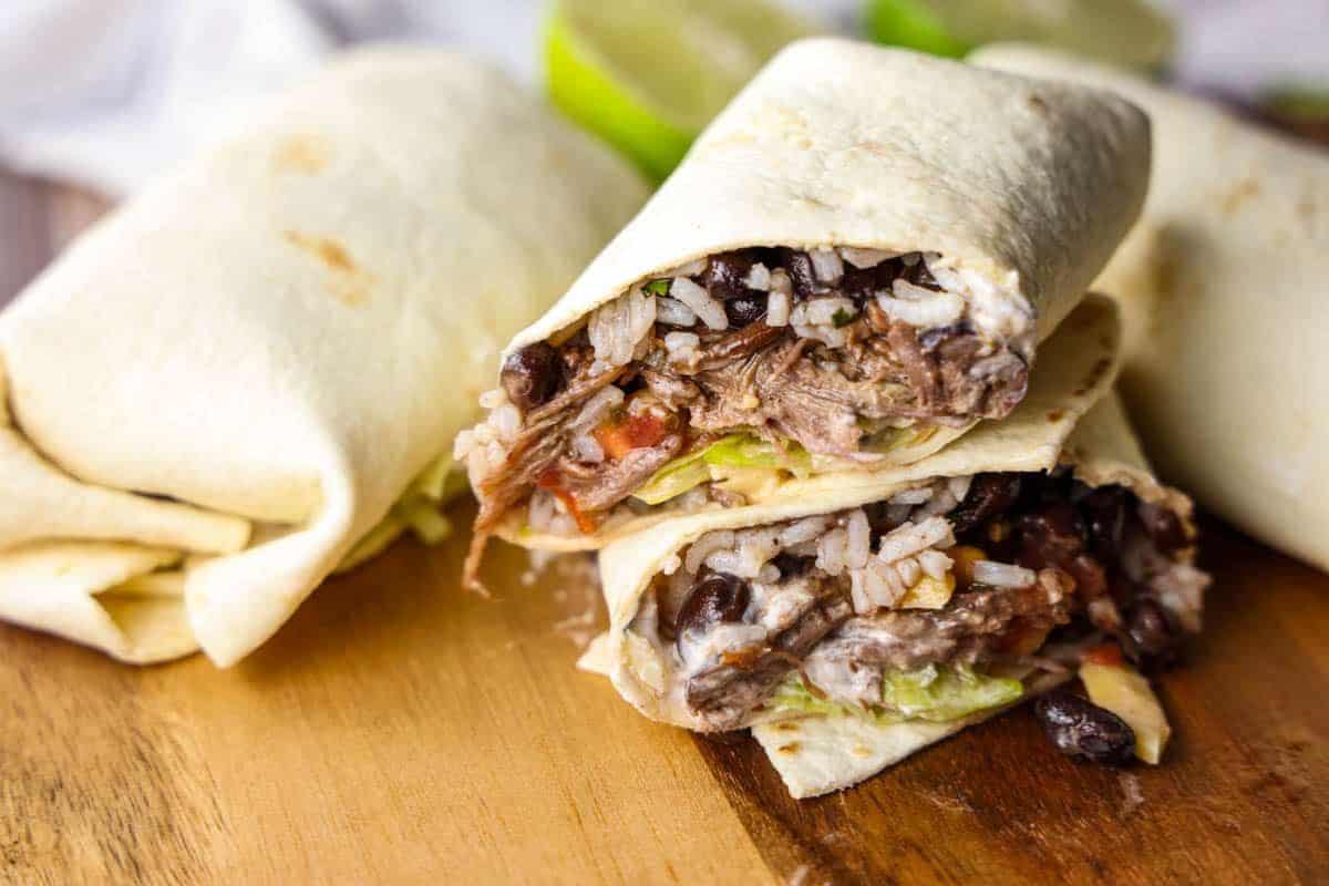 Close-up of a homemade burrito cut in half, revealing fillings of shredded meat, black beans, rice, tomatoes, and lettuce on a wooden surface. Lime wedges are visible in the background—a perfect dish for Cinco de Mayo celebrations.