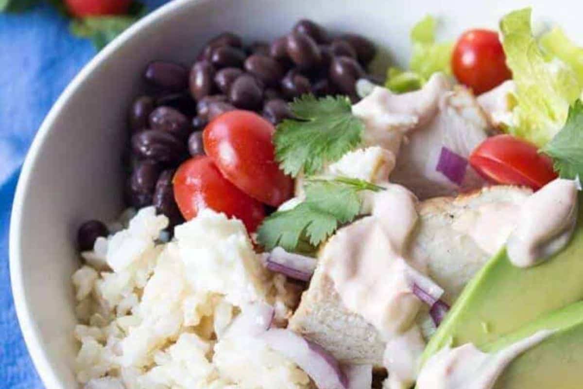 A vibrant bowl perfect for Cinco de Mayo, featuring rice, black beans, cherry tomatoes, cilantro, and avocado. Topped with sliced chicken, red onions, and a creamy sauce, this dish brings the essence of homemade burritos to your table in every bite.