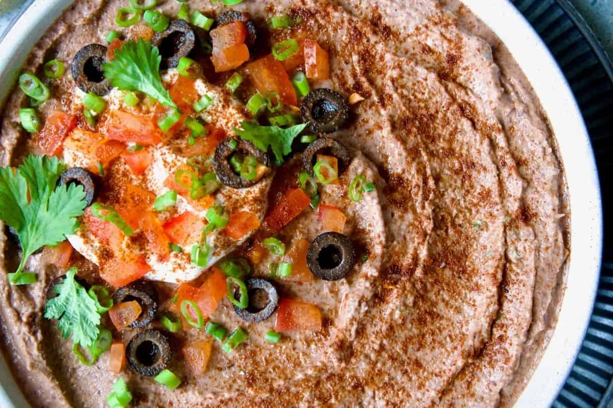 A festive bowl of creamy bean dip perfect for a Cinco de Mayo party, topped with diced tomatoes, sliced black olives, green onions, a dollop of sour cream, cilantro, and a sprinkle of paprika. A delicious dip that will be the star of any celebration.