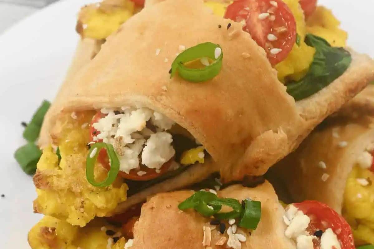Close-up of baked pastry pockets filled with scrambled eggs, sliced cherry tomatoes, green onions, and topped with crumbled cheese and sesame seeds on a white plate—perfect for your spring brunch recipes.