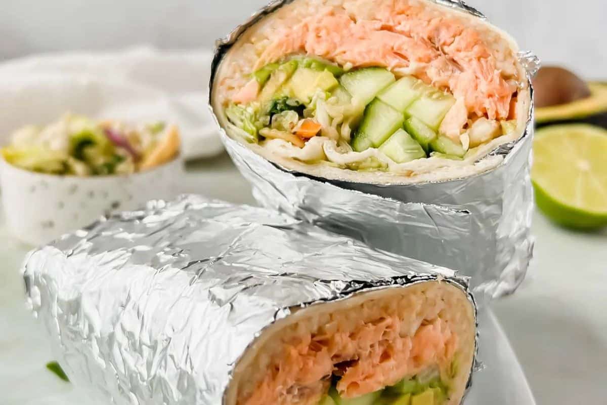 Foil-wrapped sushi burrito cut in half reveals layers of salmon, avocado, cucumber, and veggies inside a wrap—a delightful fusion perfect for Cinco de Mayo. Lime and salad adorn the background.