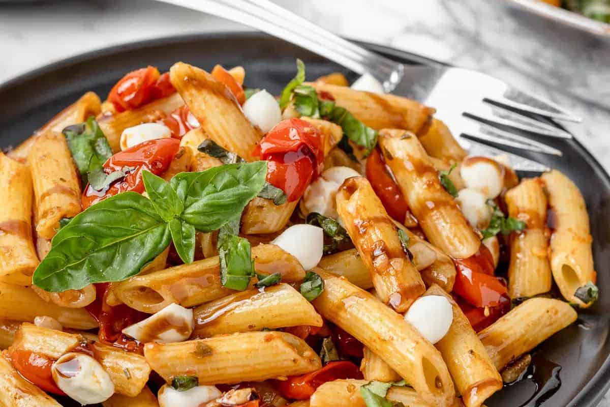 A must-try pasta dish, this plate of penne features cherry tomatoes, mozzarella balls, and fresh basil. Perfect for spring, it's artfully served on a black dish with a fork.