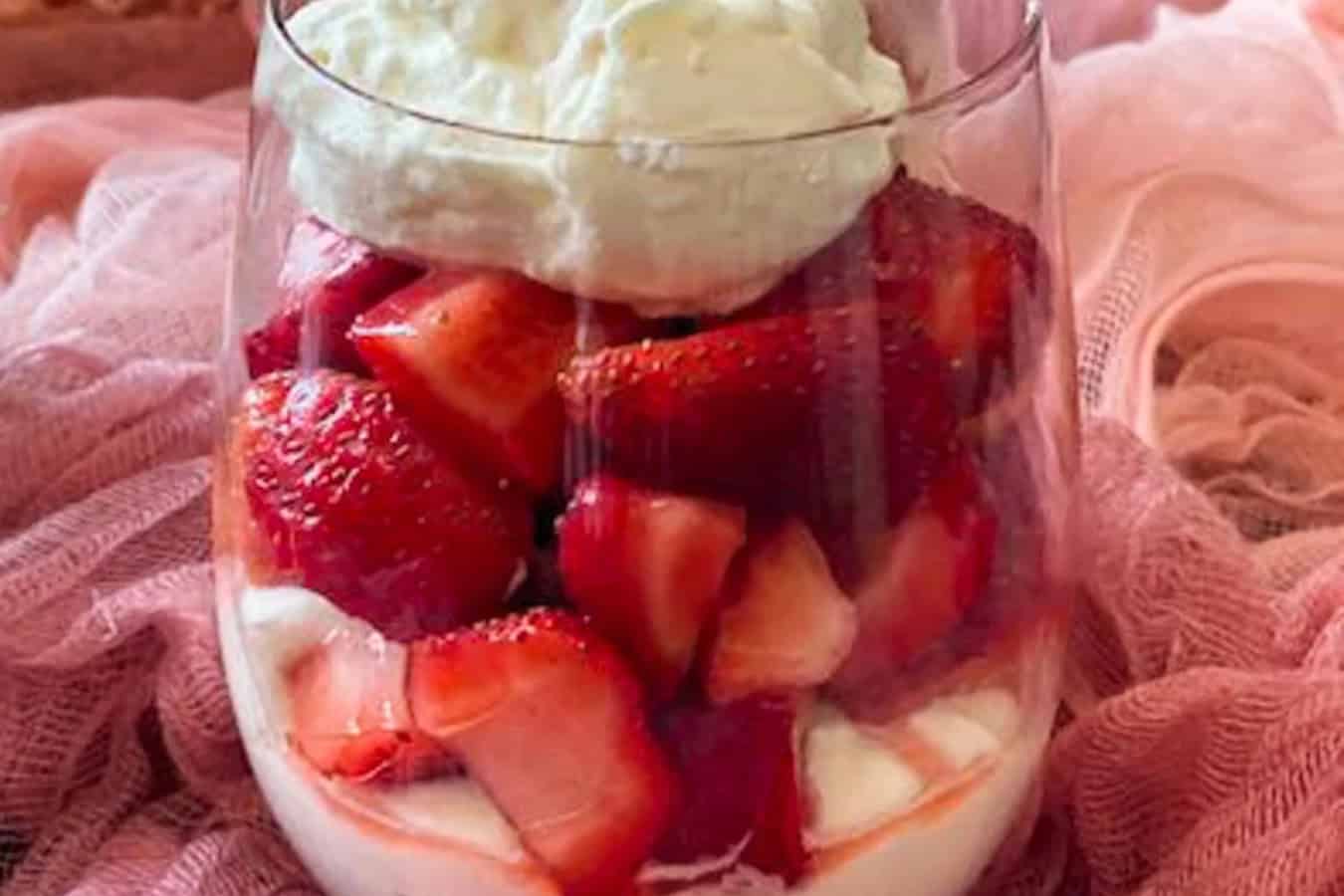 A glass showcasing a beautiful dessert of layered sliced strawberries with cream on the bottom and top, resting delicately on a pink fabric backdrop.