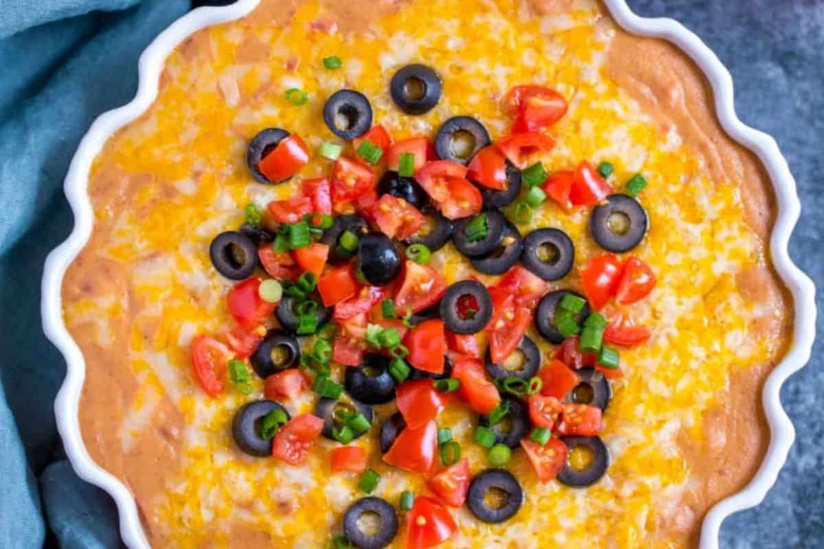 A festive cheesy rotel bean dip in a scalloped dish, perfect for a Cinco de Mayo party. It's topped with chopped tomatoes, black olives, green onions, and melted cheese to delight your guests.