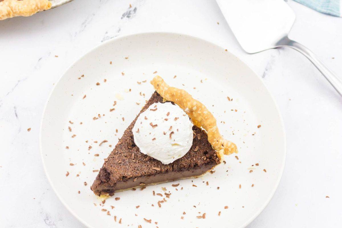 A slice of chocolate pie topped with a dollop of whipped cream on a white plate, sprinkled with chocolate shavings, sits next to a pie server—a decadent addition to any Cinco de Mayo dessert lineup.