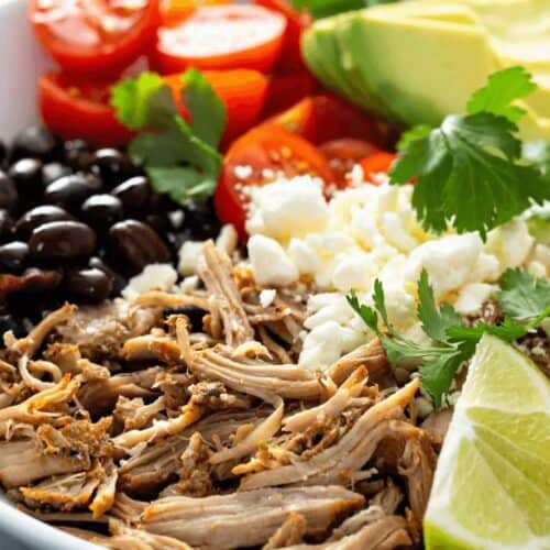 A vibrant bowl perfect for Cinco de Mayo, featuring shredded meat, black beans, cherry tomatoes, sliced avocado, cilantro, crumbled cheese, and lime wedges.