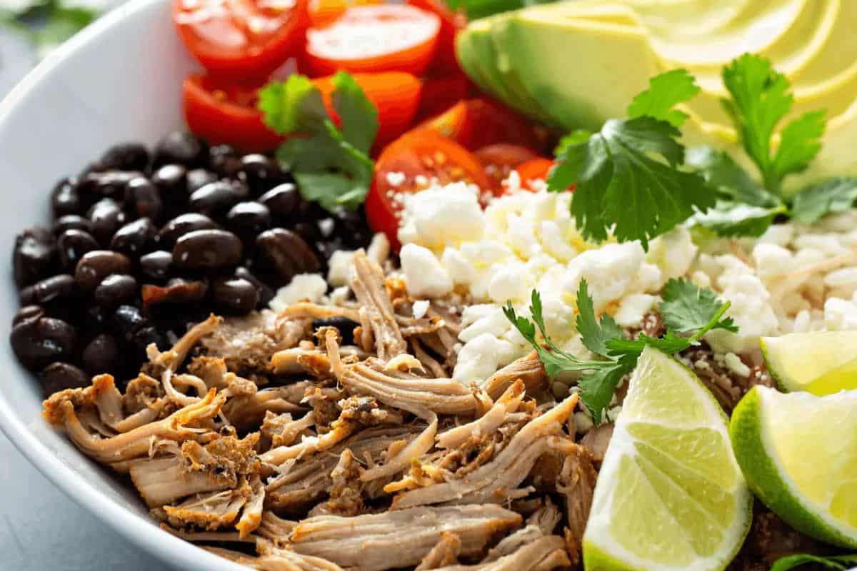A vibrant bowl perfect for Cinco de Mayo, featuring shredded meat, black beans, cherry tomatoes, sliced avocado, cilantro, crumbled cheese, and lime wedges.