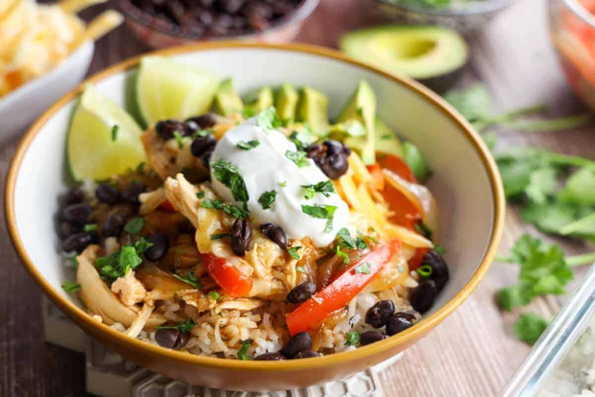 A festive bowl perfect for Cinco de Mayo, featuring rice layered with juicy chicken, black beans, sliced avocado, red peppers, shredded cheese, a dollop of sour cream, and cilantro. Lime wedges add zing to this colorful celebration.