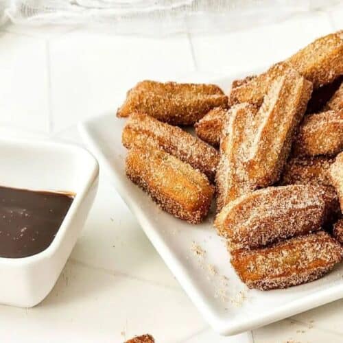 A plate of sugar-coated churros is piled next to a small square dish filled with chocolate sauce on a white surface, making for the perfect fiesta desserts.