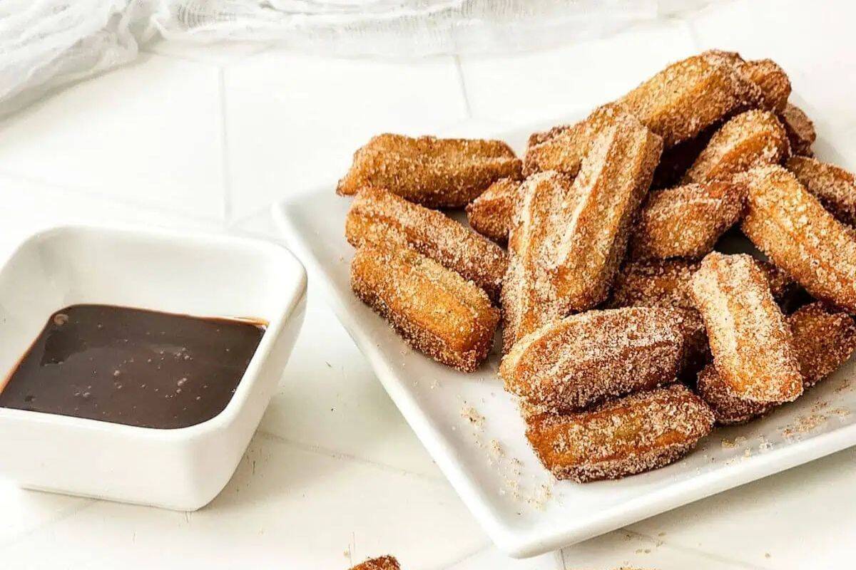 A plate of sugar-coated churros is piled next to a small square dish filled with chocolate sauce on a white surface, making for the perfect fiesta desserts.