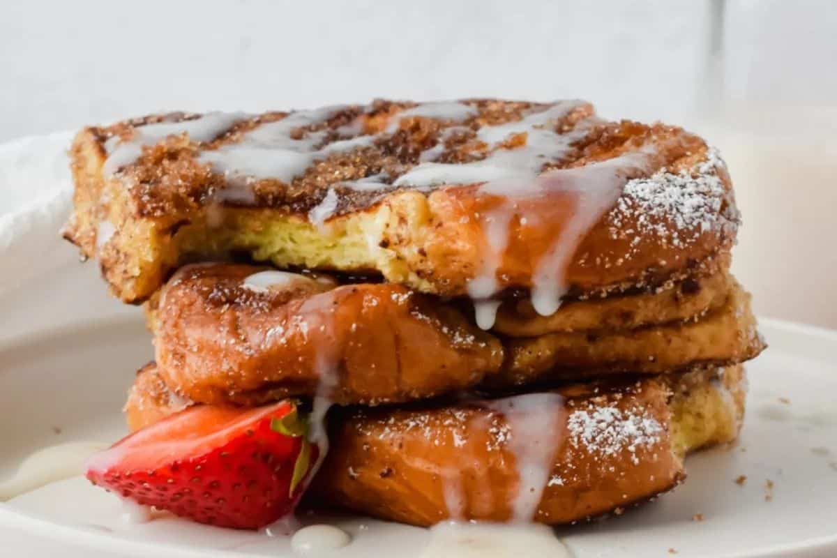 A delightful stack of churro French toast, perfect for Easter brunch, is topped with powdered sugar and icing, garnished with a strawberry slice on a pristine white plate.