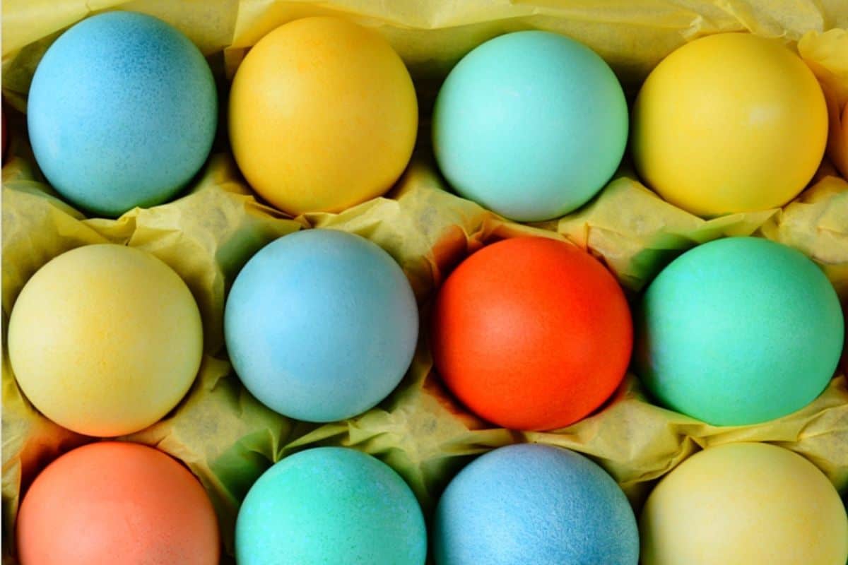 A carton cradles twelve pastel-colored eggs, arranged in four rows, with hues of blue, green, yellow, and one bright red egg. Nestled against a background of yellow packing material, it's a perfect setup for Easter egg decorating that inspires creative ideas.