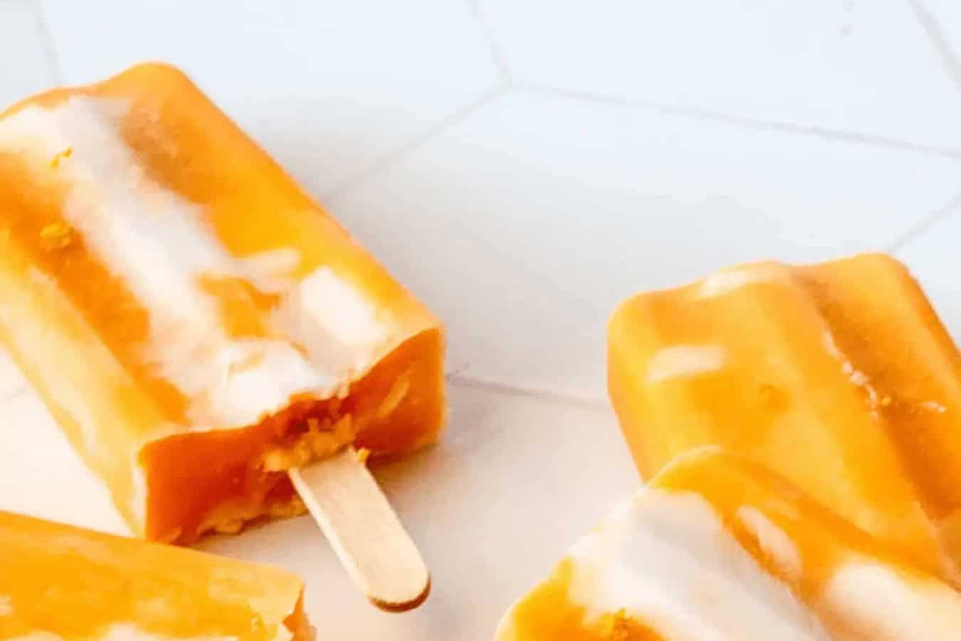 Orange and white popsicles rest on a white hexagonal tiled surface; one has a bite taken out of it, perfectly capturing the essence of beautiful spring desserts that taste as good as they look.
