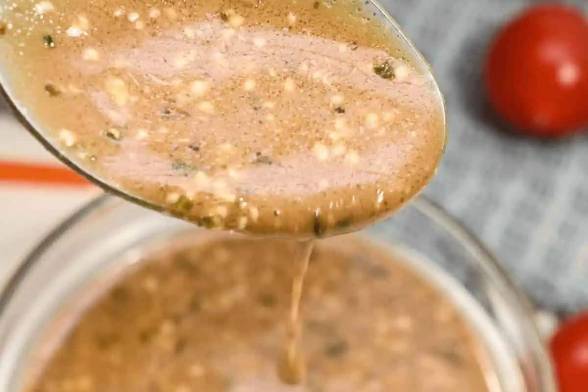Creamy balsamic dressing with visible herbs and spices being poured from a spoon into a clear glass bowl.