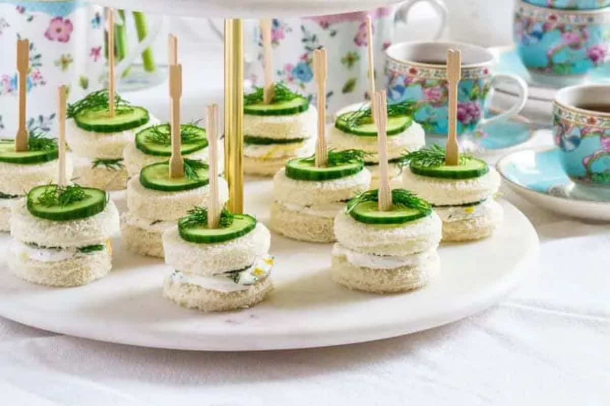 Round cucumber sandwiches on sticks are adorably arranged on a white plate, surrounded by floral-patterned teacups and teapots—perfect for your baby shower recipes collection.