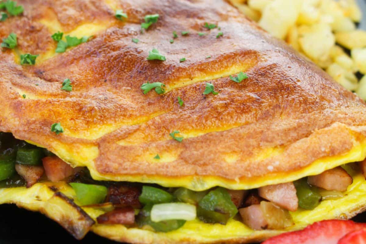 Close-up of a folded omelette filled with diced green peppers and meat, garnished with parsley, accompanied by a side of scrambled eggs. Perfect for your Easter brunch recipes collection.
