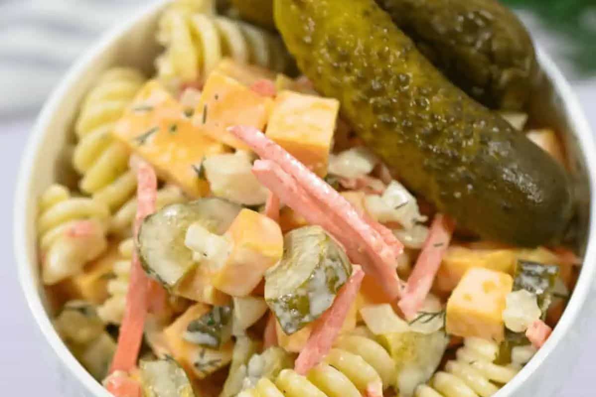 A bowl of pasta salad with rotini, diced cheese, carrots, pickles, and a creamy dressing is crowned with whole pickles—truly one of the best spring salads for the season.