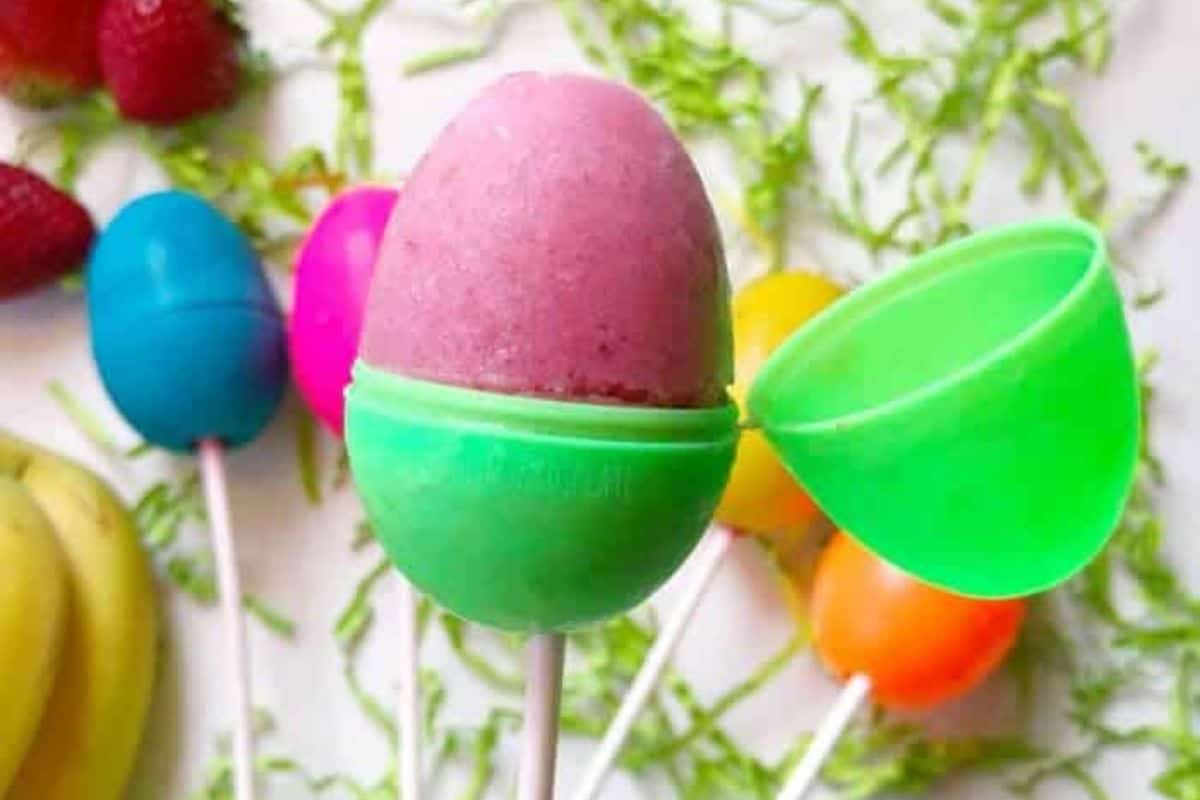Colorful frozen treats in egg-shaped molds on sticks make for creative Easter 2023 snacks. A green mold is open, revealing a pink delight. In the background, bananas and strawberries rest on vibrant green shredded paper, adding to the festive Easter egg decorating atmosphere.