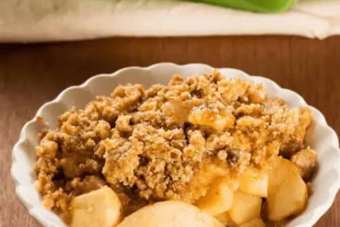 A white bowl of apple crumble with a crumbly topping rests on a wooden surface, showcasing one of the most beautiful desserts.